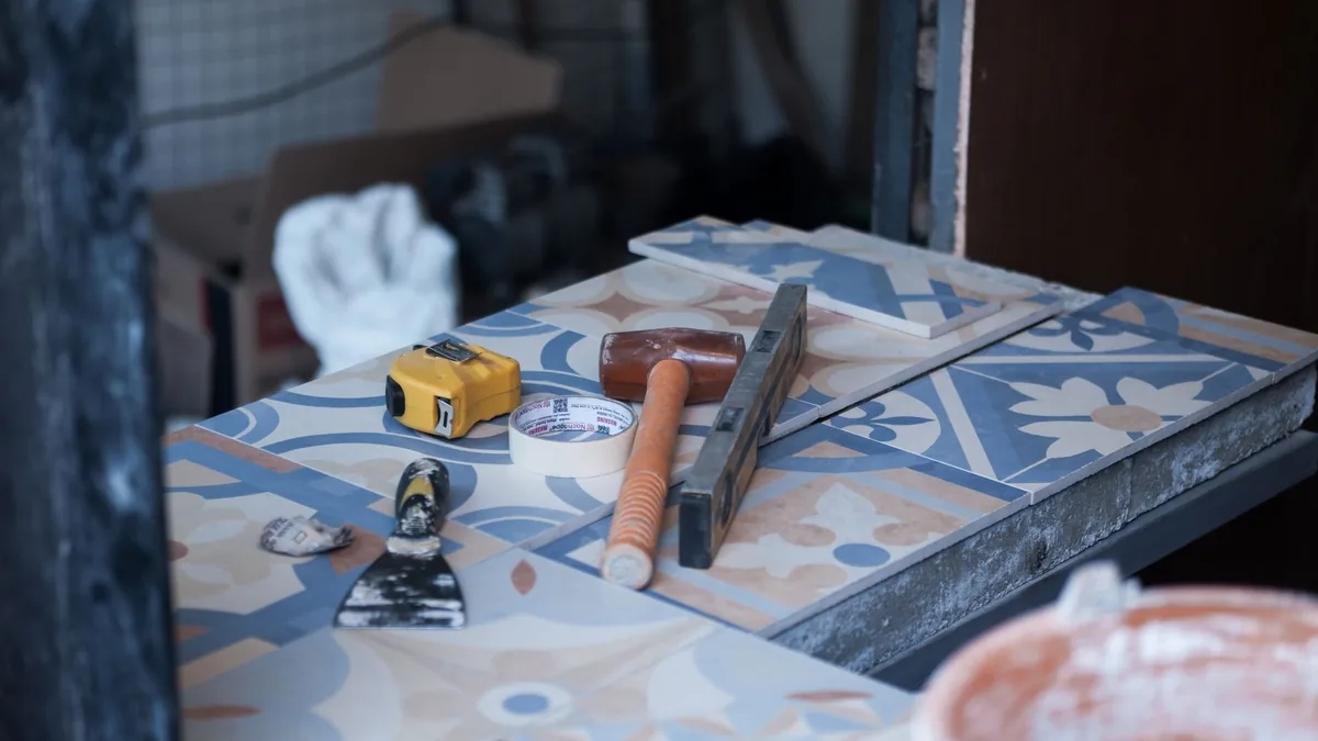 Baldosas con dibujo azul y beige sobre una mesa de obra, con un metro, un rollo de cinta adhesiva, un mazo, un nivel y una espátula con restos de cemento.