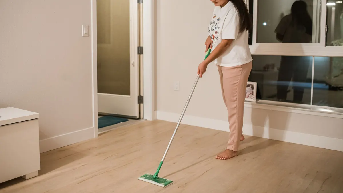 Una mujer pasa una mopa plana por un suelo de lamas en tono madera clara, en una habitación casi vacía.