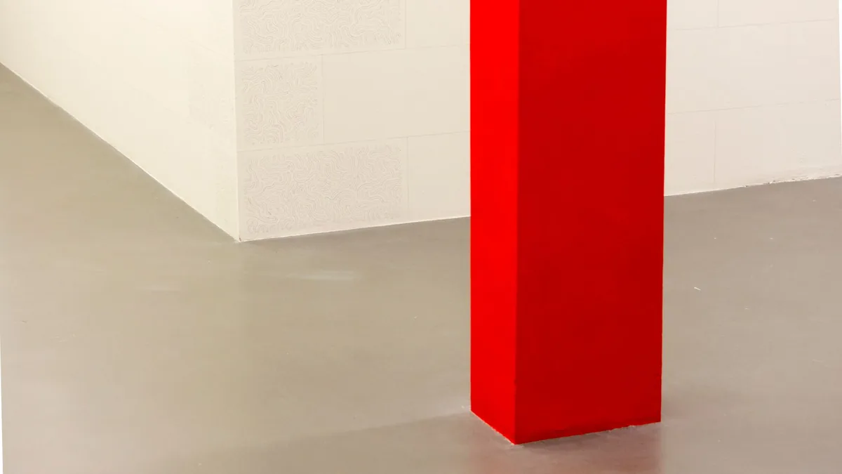 Suelo gris liso y continuo, sin juntas a la vista, junto a una pared blanca y una columna pintada de rojo.