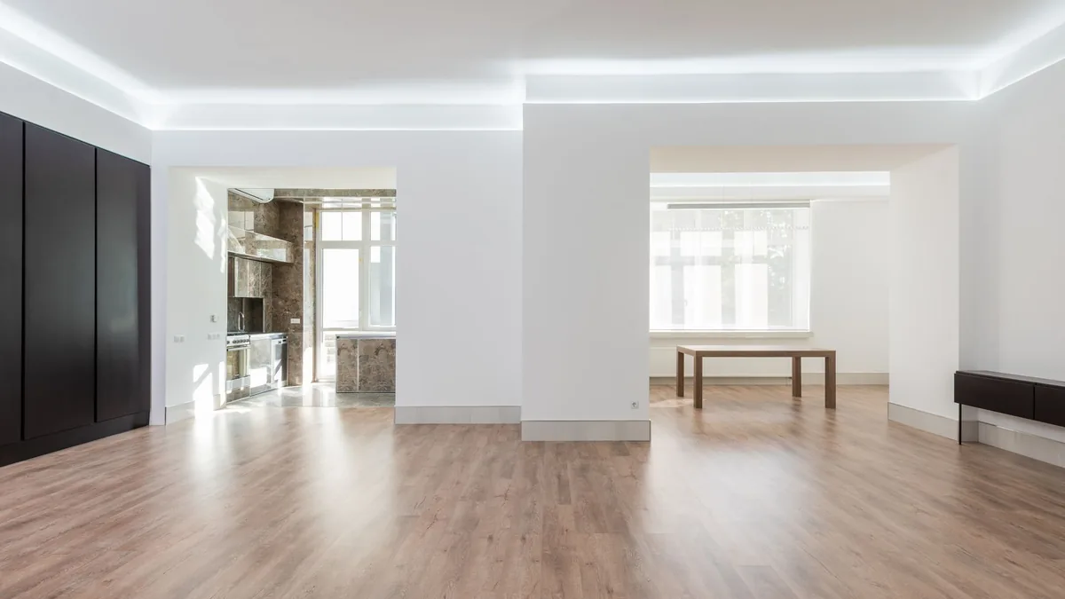 Salón luminoso con suelo de madera visto en profundidad, con la luz entrando por un ventanal lateral.